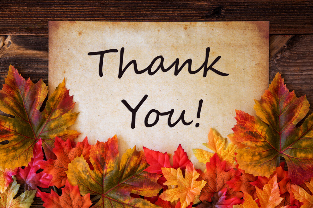 An Image of a white card and fall colored leaves on a drak stained wood table. The white card says "THANK YOU"
